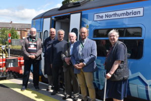 6 SENRUG Committee Members stand in front of a train at Ashington station. The train is branded in special Northumberland Line livery and is named "The Northumbrian".