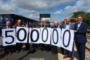 A group of people including SENRUG Chair Dennis Fancett holding a sign saying five hundred thousand in front of a Northumberland Line train at Ashington station