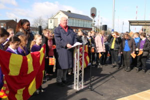 Dennis Fancett, Chair of SENRUG is standing at a lectern and speaking, with school children holding signs and waving flags in the background.